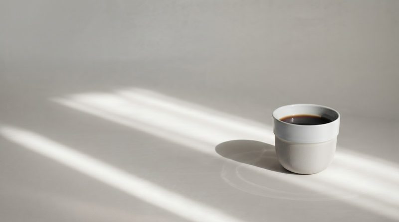 A single white ceramic cup of black specialty coffee on a pale surface, cut across by hard window light.