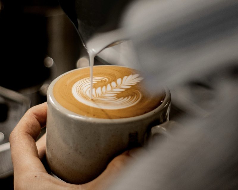 A batchhouse barista pouring textured milk into a stoneware cup, forming a rosetta on the crema.