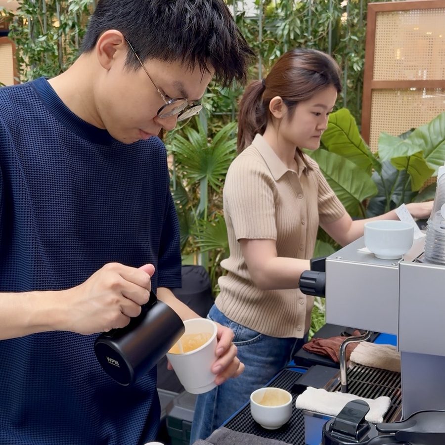 Two batchhouse baristas working side by side at the mobile coffee bar at an event — one pouring steamed milk, one at the espresso machine, framed by greenery.